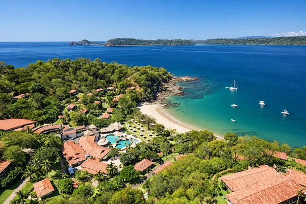 Aerial view of a beachfront resort with red-tiled roofs, pool, cove beach and boats anchored offshore