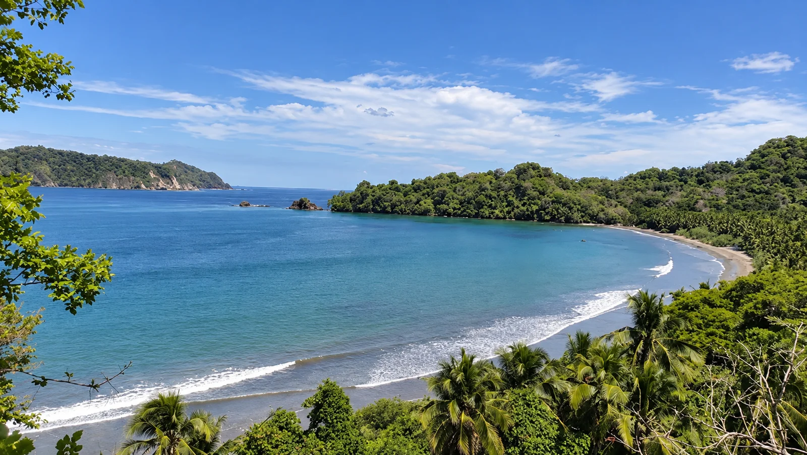 A forested coastline curves around a calm turquoise bay with a sandy beach and small rock outcrops