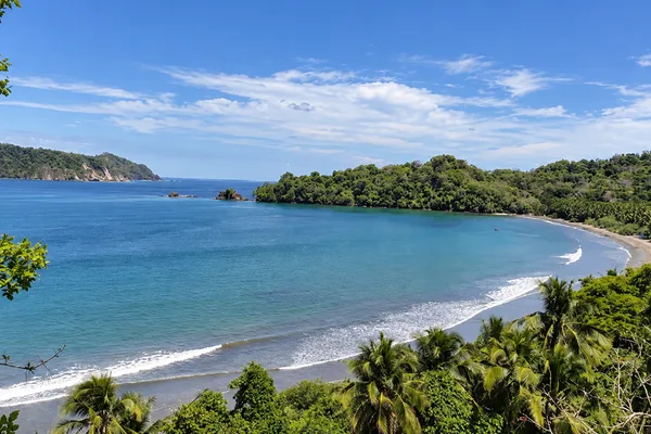 A forested coastline curves around a calm turquoise bay with a sandy beach and small rock outcrops