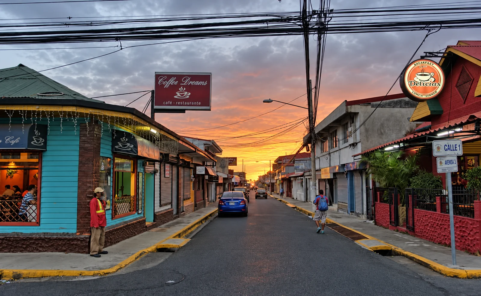 Quiet town street at sunset with cafés, a blue car, a pedestrian and power lines under an orange and pink sky