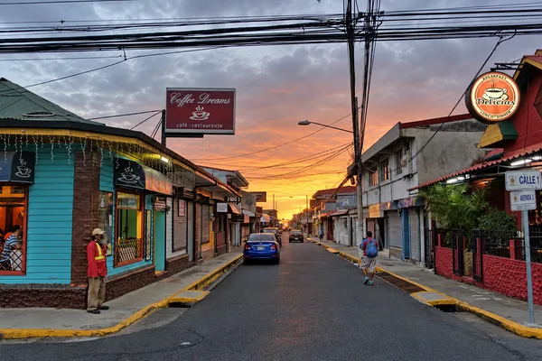 Quiet town street at sunset with cafés, a blue car, a pedestrian and power lines under an orange and pink sky